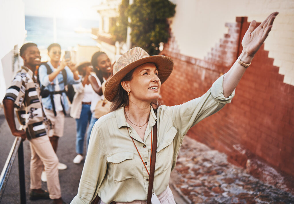 A woman in a straw hat leads a small group of travelers through a narrow sun-lit street, gesturing toward a point of interest ahead.