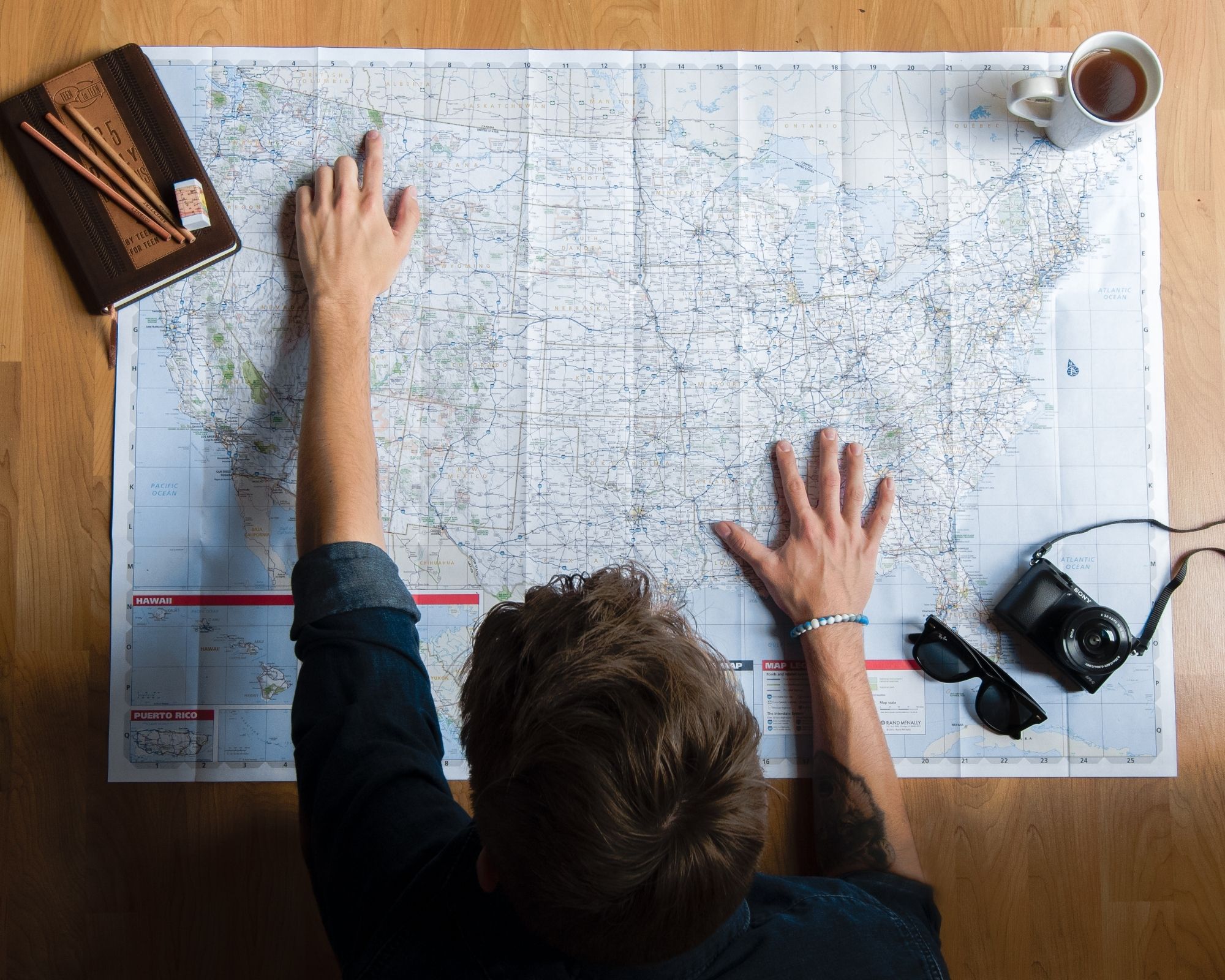 A US road map spread across a wooden table, with hands resting on it as someone leans over to study it, alongside a notebook, pencils, a camera, and a cup of tea