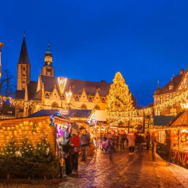 A European Christmas market at dusk, wooden stalls strung with lights beneath timbered buildings and a tall lit tree, cobblestones wet underfoot
