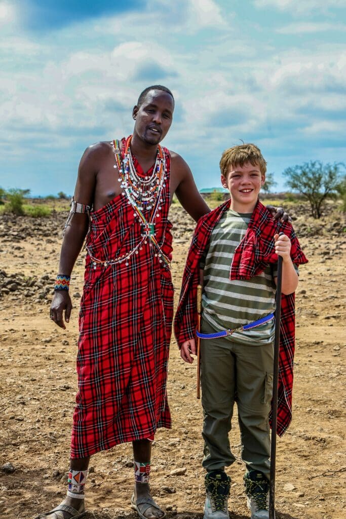 A boy wrapped in a red Maasai shuka standing with a Maasai warrior in traditional beaded dress on the Kenyan savanna.