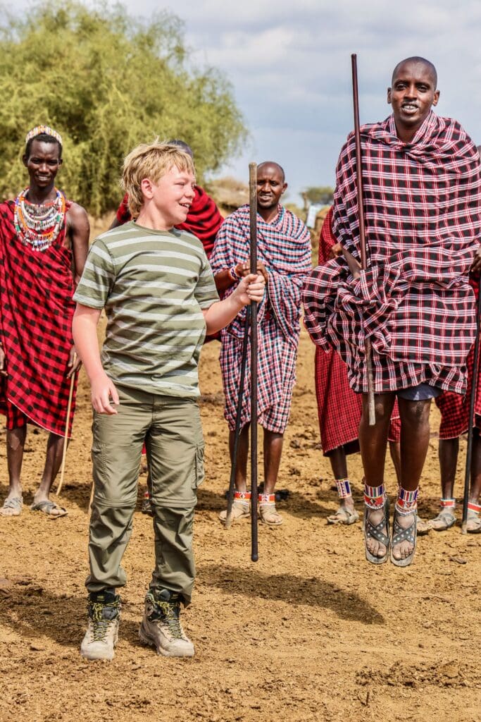 A boy laughing and holding a stick as Maasai warriors in red shukas perform their traditional adumu jumping dance in Kenya.